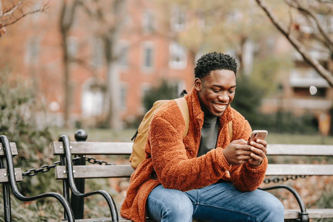A person looking happy, sitting on an outdoor bench, using their phone